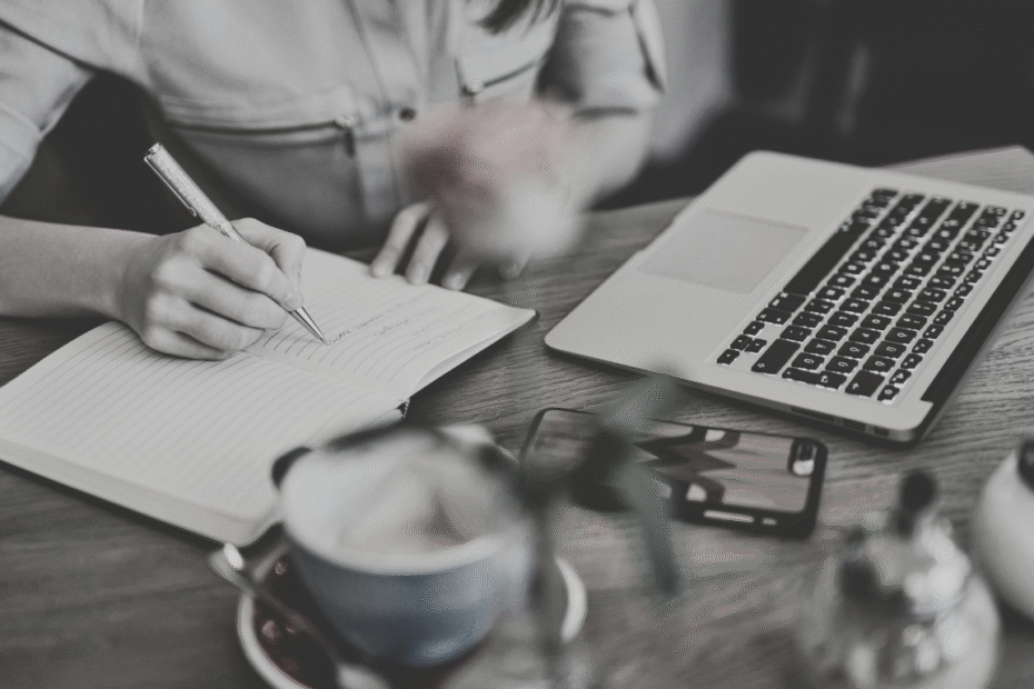 Mulher sentada em uma mesa com um computador, uma xícara, um celular e escrevendo em um caderno planejando uma estratégia de marketing de conteúdo.