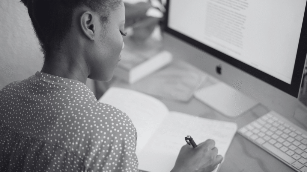Mulher escrevendo em um caderno enquanto aprende como escrever um artigo de blog, com a tela do computador ao fundo.