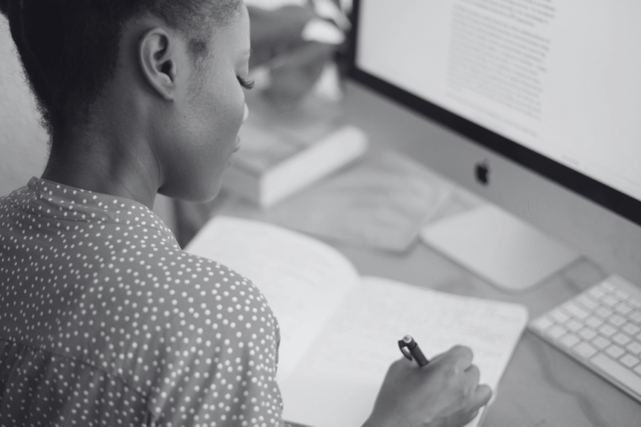 Mulher escrevendo em um caderno enquanto aprende como escrever um artigo de blog, com a tela do computador ao fundo.